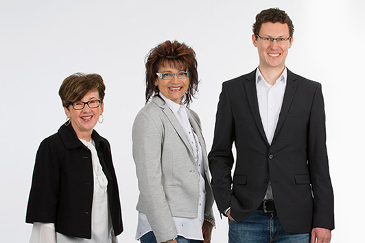 Drei Personen in Businesskleidung stehen lächelnd vor weißem Hintergrund für ein Teamfoto.